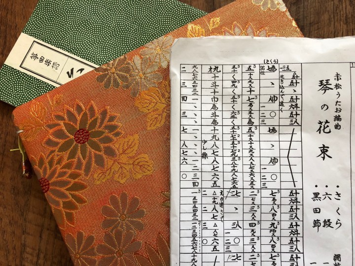 A piece of koto music -- Japanese characters printed in boxes on white paper -- sits on top of a orange and floral brocade case, which sits on top of the green cover of a piece of koto sheet music.