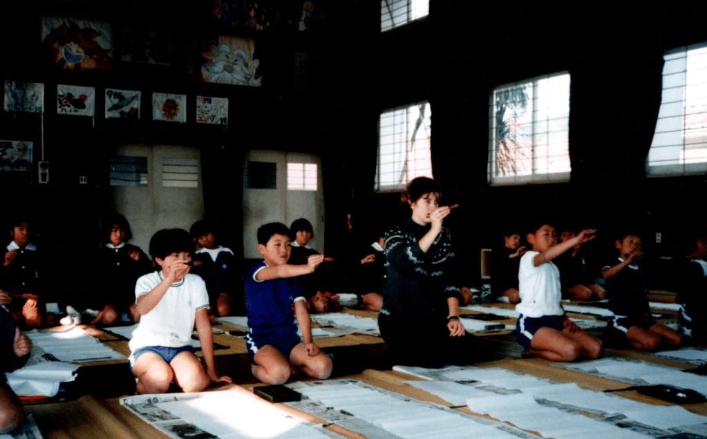 A woman sits on the floor among children, everyone holding a calligraphy brush in the air
