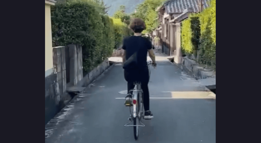 a woman bicycles down a narrow street past tile-roofed Japanese homes