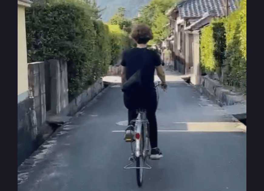 a woman bicycles down a narrow street past tile-roofed Japanese homes